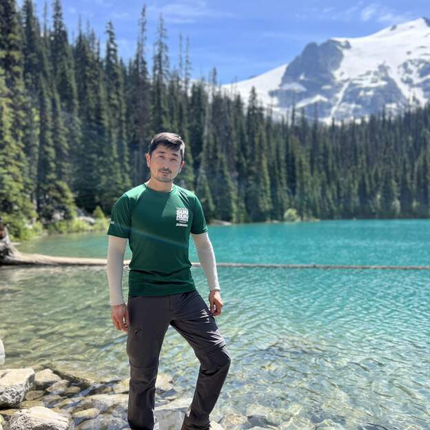 Logan stands in front of a pristine blue lake bordered by evergreen trees. Behind him in the background is a big white mountain. Logan is wearing a green t-shirt and black pants. He is posed in front of the scenery. 